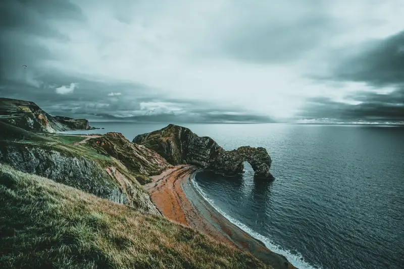 A rocky sea arch beside an orange shoreline and teal water beneath a cloudy sky.